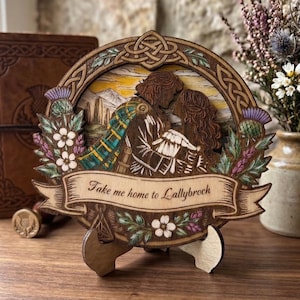 Può includere: Elemento decorativo in legno raffigurante una coppia che si bacia, con la scritta "Take me home to Lallybroch". Il design include elementi floreali e di cardo, e un nodo celtico. Il pezzo è esposto su un supporto in legno.