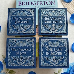 Peut inclure: Quatre dessous de verre carrés bleu marine avec du texte et des motifs argentés, chacun avec un titre de la série de livres "Bridgerton". Un livre bleu clair et des pétales de fleurs bleues entourent les dessous de verre.