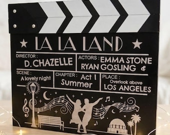 May include: A black and white film clapperboard with the text "LA LA LAND". The clapperboard features the director, actors, scene, chapter, and place. The bottom of the clapperboard has a graphic of a couple dancing, a bench, and musical notes.