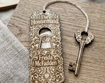 May include: A wooden bookmark with the text "The Housemaid Freida McFadden" and an ornate keyhole design. A decorative key is attached with a string. The bookmark and key are made of light-colored wood.