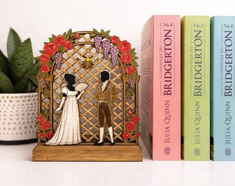 May include: Wooden bookend featuring a laser-cut design of a couple in period attire, a bee, and floral accents. Three books with the title "Bridgerton" stand beside the bookend. The books are in pink, green, and blue.