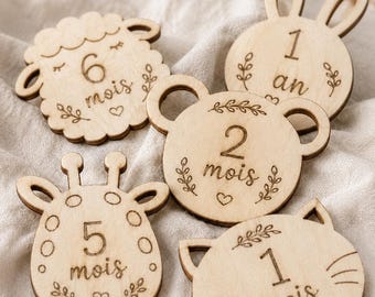 May include: Five light-colored wooden milestone discs featuring animal shapes and engraved text. The discs include a sheep labeled 6 mois, a rabbit labeled 1 an, a bear labeled 2 mois, a giraffe labeled 5 mois, and a cat labeled 1 mois, all decorated with small leaf and heart motifs.