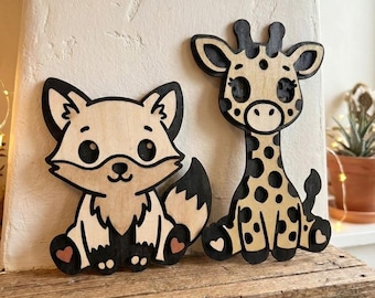 May include: Two wooden animal cutouts: a fox and a giraffe. The fox is light brown with black accents and a heart detail. The giraffe is light brown with black spots and accents. Both are sitting on a wooden shelf.