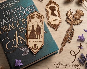 May include: Wooden bookmarks with intricate designs, including a silhouette, a couple, and a thistle. The bookmarks are placed on a teal book with the title "Dragon in Amber" and the word "Outlander".