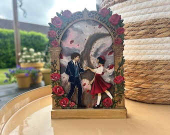 May include: A wooden decorative piece featuring a couple dancing under a rose-covered arch. The man wears a blue suit, and the woman is in a red dress. The background depicts a swirling, abstract scene. Red roses adorn the frame.