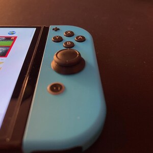 Könnte beinhalten: Nahaufnahme eines türkisfarbenen Gamecontrollers mit schwarzen Knöpfen und einem Joystick. Der Bildschirm zeigt farbenfrohe Spielgrafiken. Der Controller befindet sich auf einer dunklen Oberfläche, was auf ein Gaming-Setup hindeutet.