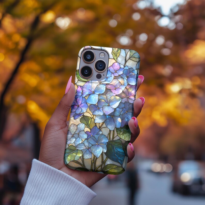 May include: A smartphone case featuring a stained-glass design of blue and purple hydrangea flowers with green leaves. The case has a glossy finish and a protective design. The case is held in a hand with pink nail polish.