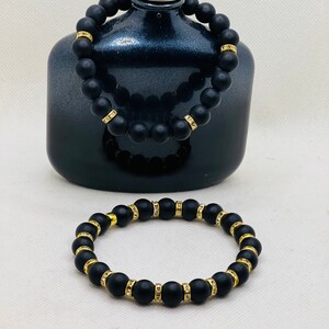 May include: Two black beaded bracelets with gold accents and clear rhinestones. The bracelets are on a white surface.
