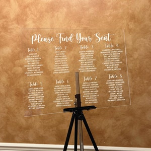 May include: A clear acrylic sign with the text "Please Find Your Seat" at the top. The sign is divided into eight sections, each labeled with a table number and a list of names. The sign is standing on a black tripod.