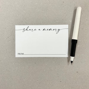 May include: A white card with the words "share a memory" in elegant script. Below the text is a line with the word "FROM:" printed on the card. A white pen with a black grip rests on the right side of the card.