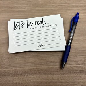 May include: White card with black text that reads "Let's be real... Advice for the mom to be" with blank lines for writing and the word "love," followed by a blank line at the bottom. A blue pen is lying on a wood surface next to the card.