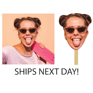 May include: A cutout of a woman's face with brown hair and sunglasses. The cutout is attached to a stick. The woman is sticking her tongue out and smiling. The background is a light orange color.