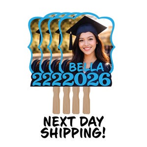 May include: Graduation photo booth prop featuring a young woman in a graduation cap. The prop has a blue scalloped border with the name "BELLA" and the year "2026" in blue. The prop is attached to a wooden stick.