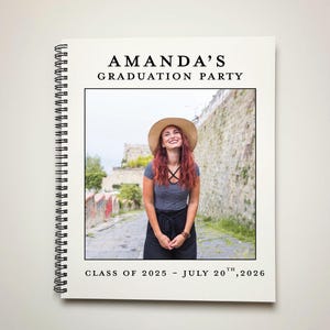 May include: A white graduation party invitation with a black border. The invitation has a photo of a woman wearing a hat and smiling. The text on the invitation reads "Amanda's Graduation Party, Class of 2025 - July 20th, 2026".