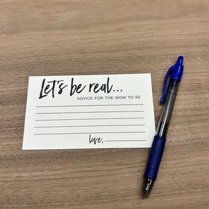May include: A white card with black text that reads "Let's be real... Advice for the mom to be Love," with blank lines for writing. A blue pen is lying on a wooden surface next to the card.