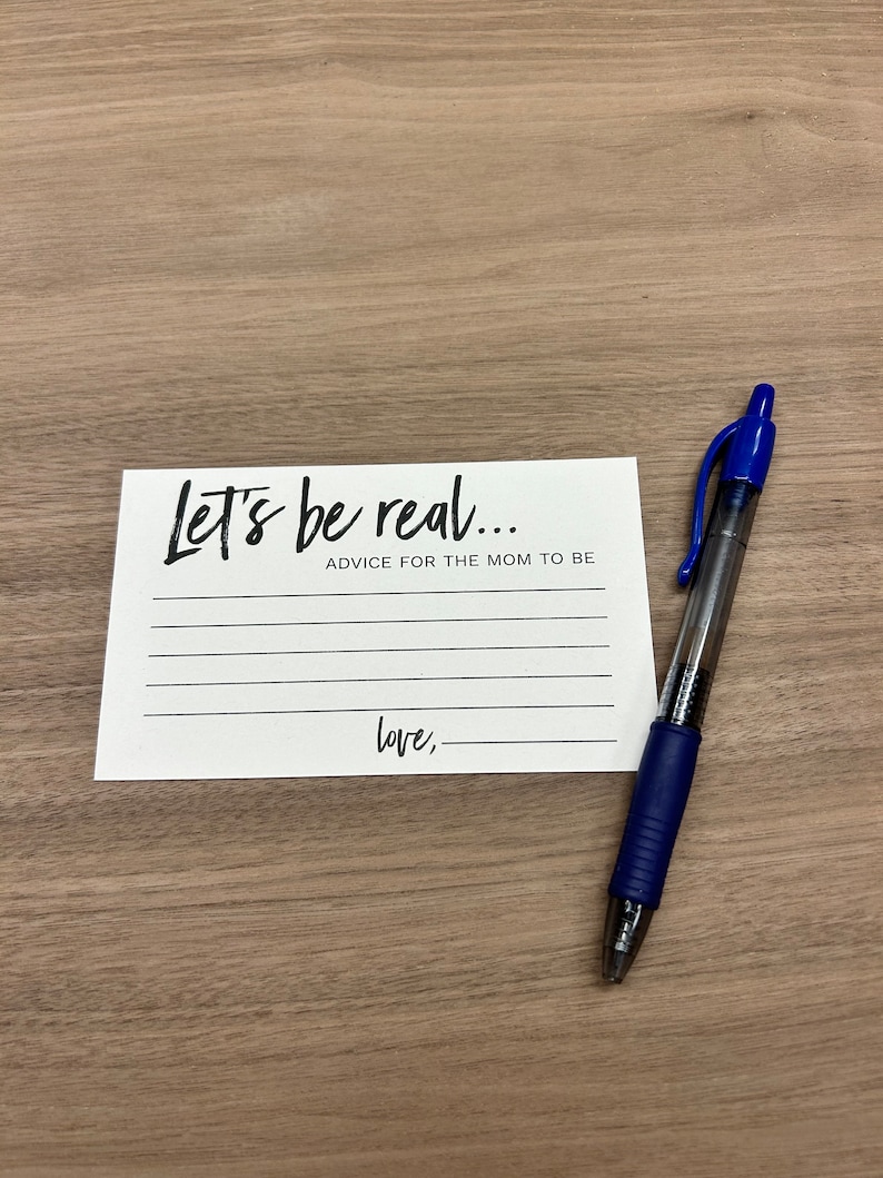 May include: A white card with black text that reads "Let's be real... Advice for the mom to be Love," with blank lines for writing. A blue pen is lying on a wooden surface next to the card.
