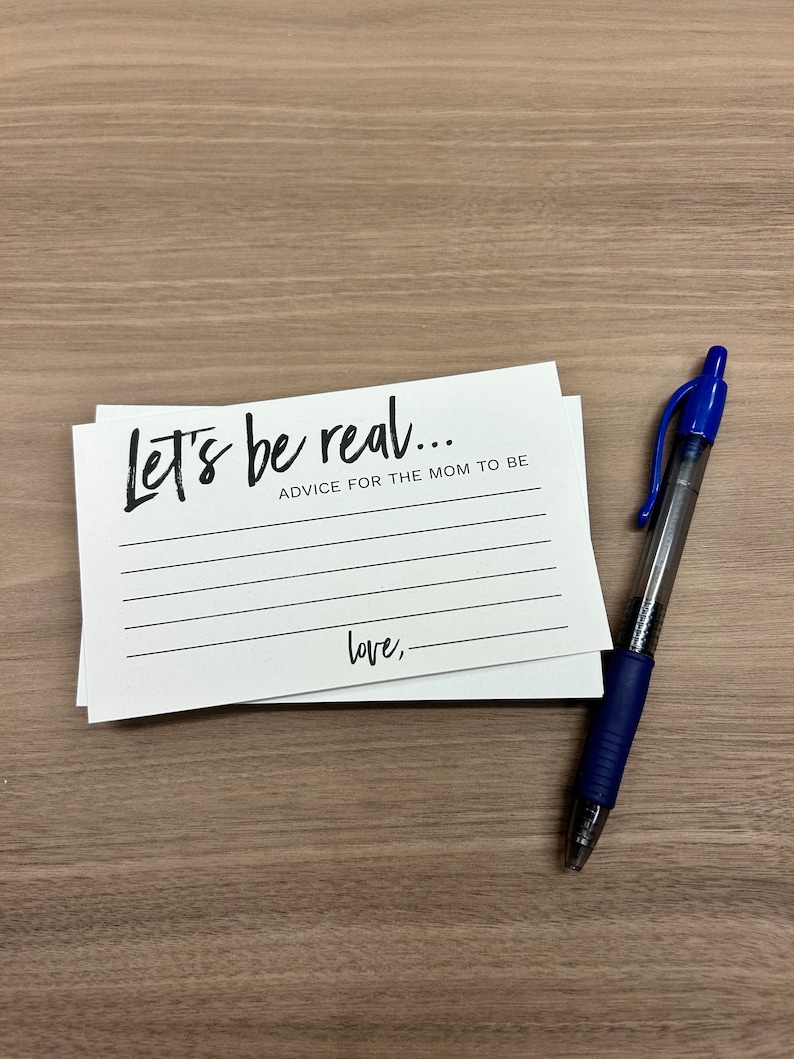 May include: White card with black text that reads "Let's be real... Advice for the mom to be" with blank lines for writing and the word "love," followed by a blank line at the bottom. A blue pen is lying on a wood surface next to the card.