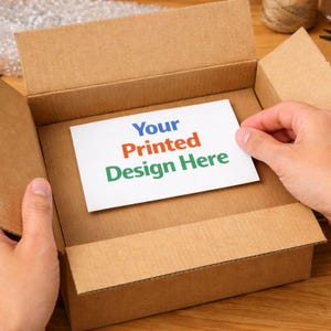 May include: A brown cardboard box with the top flaps open, revealing a white card inside. The card has the text "Your Printed Design Here" in blue, red, and green. The box is being held open by two hands.