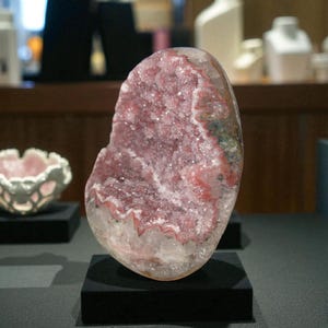 Pode incluir: Uma geodo grande e polida com um interior cristalino. A geodo apresenta uma vibrante colora&ccedil;&atilde;o rosa e vermelha dentro de um exterior transparente, semelhante ao quartzo. &Eacute; exibida em um pequeno pedestal preto.