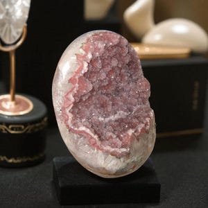 Pode incluir: Geodo oval polido, com exterior branco transl&uacute;cido e interior cristalino rosa-p&uacute;rpura brilhante. Apresentado em um pequeno suporte retangular preto, real&ccedil;ando sua estrutura natural e cor.