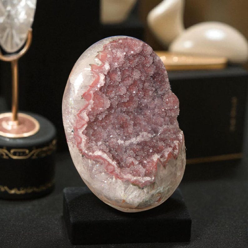 Pode incluir: Geodo oval polido, com exterior branco transl&uacute;cido e interior cristalino rosa-p&uacute;rpura brilhante. Apresentado em um pequeno suporte retangular preto, real&ccedil;ando sua estrutura natural e cor.