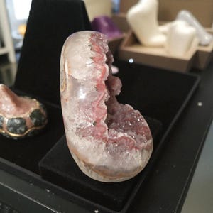 Pode incluir: Uma geodo de cristal polida, em forma oval, com um exterior transparente e um interior roxo-avermelhado vibrante. A geodo &eacute; exibida em uma superf&iacute;cie preta, mostrando sua beleza natural e intrincada estrutura cristalina. Um item decorativo &uacute;nico.