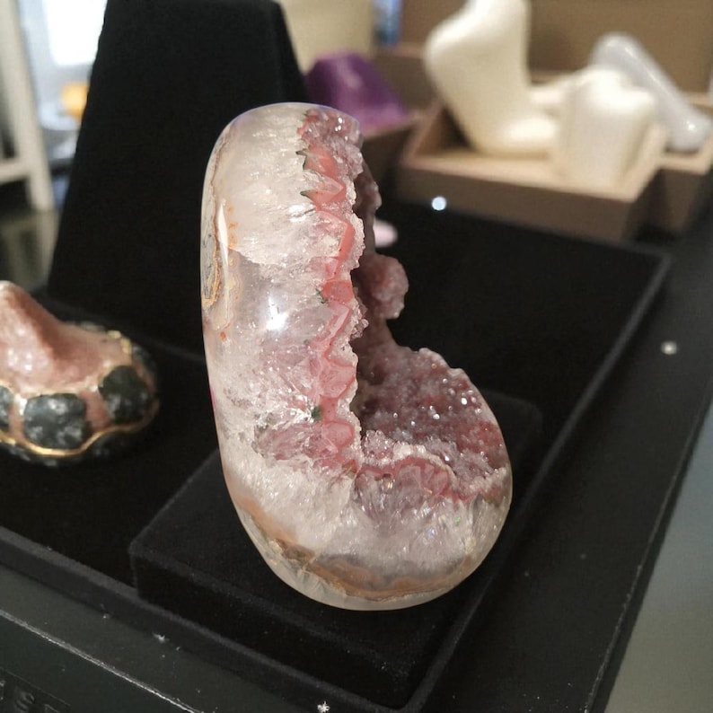 Pode incluir: Uma geodo de cristal polida, em forma oval, com um exterior transparente e um interior roxo-avermelhado vibrante. A geodo &eacute; exibida em uma superf&iacute;cie preta, mostrando sua beleza natural e intrincada estrutura cristalina. Um item decorativo &uacute;nico.