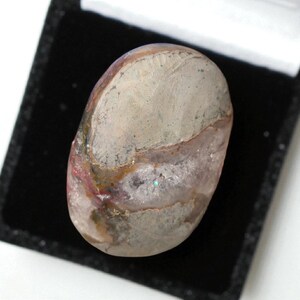 Pode incluir: Uma pedra preciosa oval polida, exibindo uma mistura de cores cinza, rosa e marrom. A pedra &eacute; apresentada numa caixa de veludo preto, real&ccedil;ando os seus padr&otilde;es naturais. A superf&iacute;cie da pedra preciosa tem uma qualidade reflexiva.
