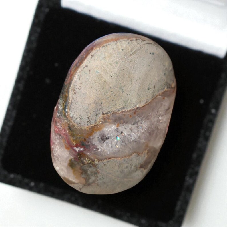 Pode incluir: Uma pedra preciosa oval polida, exibindo uma mistura de cores cinza, rosa e marrom. A pedra &eacute; apresentada numa caixa de veludo preto, real&ccedil;ando os seus padr&otilde;es naturais. A superf&iacute;cie da pedra preciosa tem uma qualidade reflexiva.