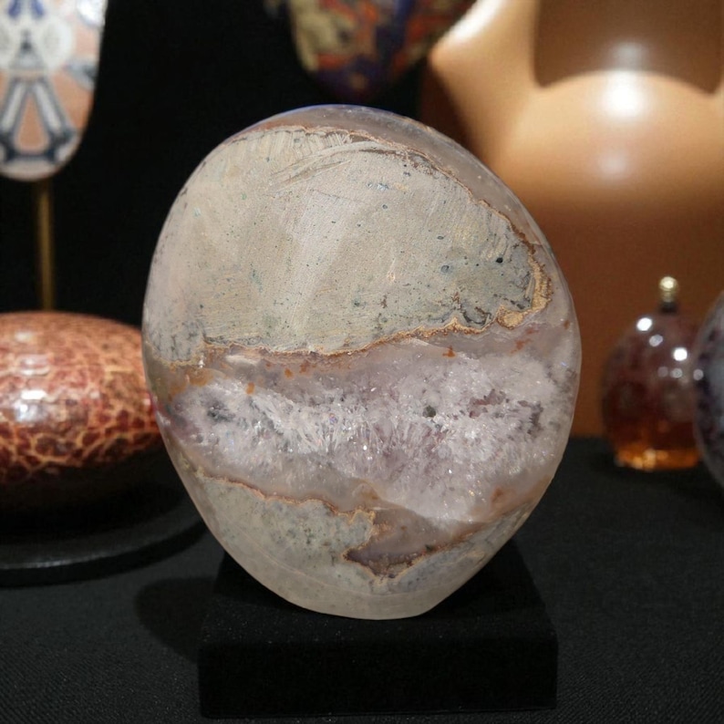 Pode incluir: Uma geodo de cristal polido, em forma oval, com uma combina&ccedil;&atilde;o de cores cinza claro e lavanda. A geodo est&aacute; sobre um pequeno suporte preto, mostrando suas texturas naturais e seu interior cristalino. A pedra tem uma parte superior lisa e arredondada e uma base &aacute;spera e texturizada.