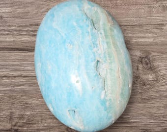 May include: A large, oval-shaped stone with a light blue hue and white veining. The stone has a smooth, polished surface and a few natural fissures. The stone is set against a wooden background.