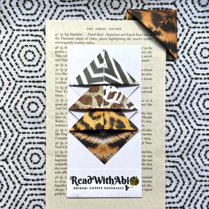 Könnte beinhalten: Origami-Eck-Lesezeichen mit Tiermustern, darunter Zebra-, Leoparden- und Tigerdesigns. Die Lesezeichen sind auf einer Seite aus "The Great Gatsby" mit dem Text "Read With Abi Origami Corner Bookmarks" abgebildet.
