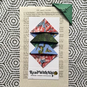 Könnte beinhalten: Origami-Eck-Lesezeichen mit Mustern wie rosa Flamingos, grünes Laub und blaue Vögel, gestapelt auf einer Seite aus "The Great Gatsby". Eine kleine Visitenkarte zeigt "Read With Abi Origami Corner Bookmarks".