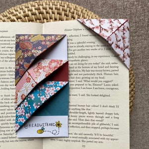 Könnte beinhalten: Origami-Lesezeichen in verschiedenen floralen und gemusterten Designs, darunter Kirschblüten und japanische Motive. Die Lesezeichen sind gefaltet und auf den Seiten eines Buches platziert. Der Text "@READWITHABI" ist sichtbar.