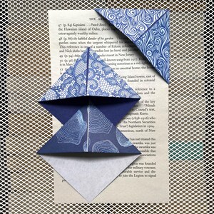 Op de afbeelding: Drie blauwe origami bladwijzers met verschillende patronen, waaronder kant, krullen en stippen, liggen op een pagina van een boek. De bladwijzers zijn gevouwen in driehoekige vormen en geven het leesmateriaal een decoratieve uitstraling.