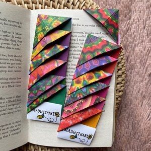Könnte beinhalten: Origami-Eck-Lesezeichen in leuchtenden Farben und Mustern, die in die Seiten eines Buches eingelegt sind. Die Lesezeichen weisen geometrische Designs in Grün, Lila, Rot und Gelb auf. Jedes Lesezeichen hat ein kleines weißes Etikett mit dem Text "READWITHABI" und einer Bienenillustration.