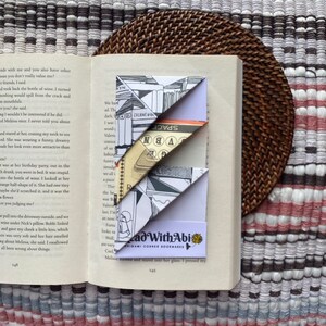 Könnte beinhalten: Ein Buch mit einem dekorativen Origami-Eck-Lesezeichen. Das Lesezeichen ist weiß mit schwarzen und grauen Illustrationen und dem Text "Read With Abi". Das Lesezeichen ist zu einer Dreiecksform gefaltet und wird auf die Ecke der Buchseiten gelegt.