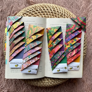 Könnte beinhalten: Offenes Buch mit farbenfrohen Origami-Eckmarkierungen. Die Lesezeichen bestehen aus gemustertem Papier in Rot-, Grün-, Gelb- und Lilatönen. Der Text "@readwithabi" und eine Bienenillustration sind sichtbar.