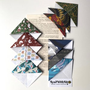 Könnte beinhalten: Eine Sammlung von Origami-Eck-Lesezeichen mit verschiedenen Mustern, darunter Weihnachtsbäume, Katzen und abstrakte Designs. Die Lesezeichen sind in die Seiten eines Buches eingelegt, mit einem "Read With Abi"-Logo.