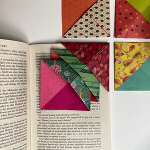 Könnte beinhalten: Bunte Origami-Lesezeichen in Dreiecksform mit verschiedenen Mustern, darunter Blätter und geometrische Designs. Die Lesezeichen liegen auf einem offenen Buch und sind in Rot-, Grün- und Orangetönen gehalten.