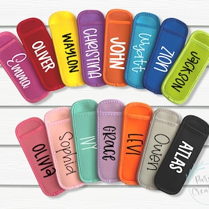 May include: A set of 14 colorful fabric popsicle holders with personalized names, including Emma, Oliver, Waylon, Christina, John, Wyatt, Zion, Jackson, Olivia, Sophia, Ivy, Grace, Levi, Owen, and Atlas.
