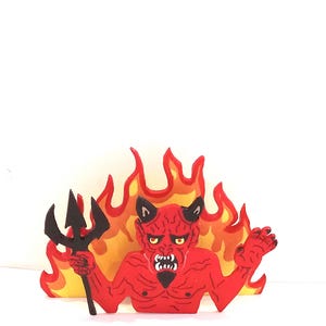 May include: A red devil figure with black horns and a black goatee, holding a black trident in one hand. The figure is positioned in front of a stylized yellow and orange flame background, featuring a fierce expression with bared white teeth.