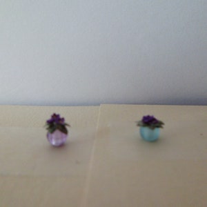 Może przedstawiać: Dwa miniaturowe doniczki, jeden jasnofioletowy, a drugi jasnoniebieski, każdy z małą, ciemnofioletową kompozycją kwiatową. Doniczki są okrągłe i wydają się być wykonane ze szkła lub podobnego materiału. Tło jest jednolite, białawe.