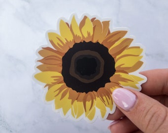 May include: A yellow sunflower sticker with a brown center. The sticker is on a white marble background.