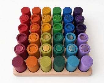 May include: A wooden tray containing thirty-six cylindrical wooden pieces arranged in a six-by-six grid. Each piece features a solid color from a rainbow spectrum, including red, orange, yellow, green, blue, and purple shades. The text Grapat is sold separately is visible at the bottom.