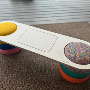 Puede incluir: Un banco de madera blanco con dos asientos redondos. Un asiento es amarillo y el otro está cubierto de confeti de colores. El banco tiene un recorte rectangular en el centro.