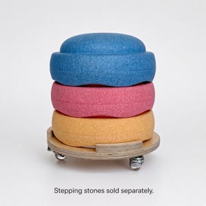 May include: A circular wooden base with four small metal caster wheels, designed to hold stacked objects. The base features a light wood grain finish and a notched rim. The text "Stepping stones sold separately" is printed below the base.