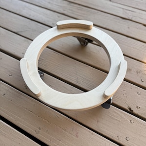 May include: A circular light-colored wooden plant caddy featuring a smooth finish and three raised curved supports along the inner rim. The base is equipped with three small black swivel casters attached to the underside for mobility.