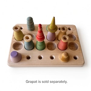 May include: A rectangular wooden board with multiple circular cutouts, featuring several small, colorful wooden figures of varying shapes and sizes placed within the holes. The text "Grapat is sold separately." is printed at the bottom.