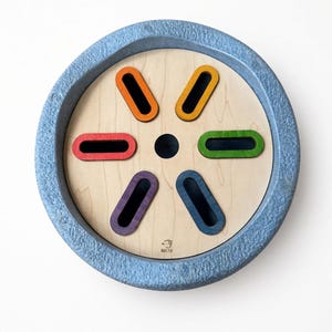 May include: A circular wooden board with a textured grey outer rim, featuring six colorful oval cutouts arranged in a radial pattern around a central black circle. The wood surface displays the Maple Play logo. Text reads: Stepping stones sold separately.